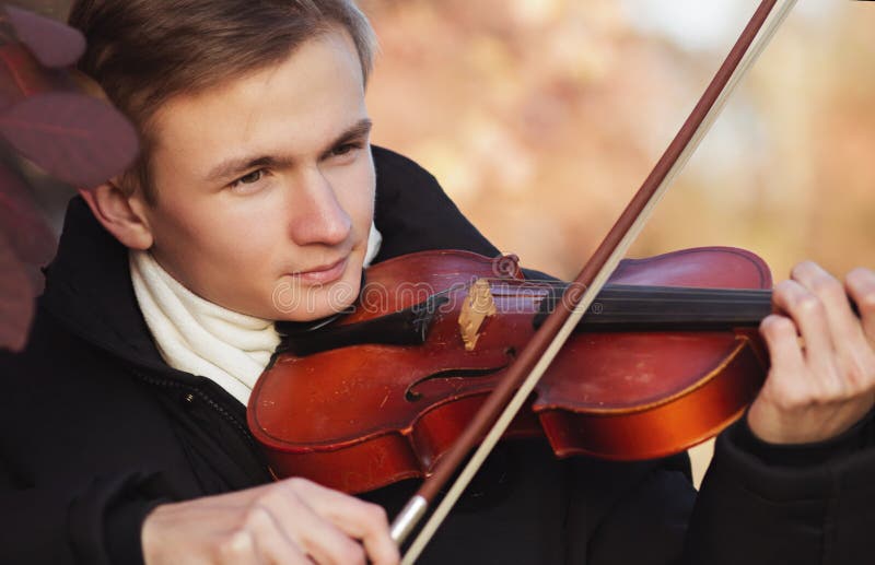 Face profile of a young elegant man playing the violin on autumn nature backgroung, a boy with a bowed orchestra instrument makes a concert, concept of classical music, hobby and art