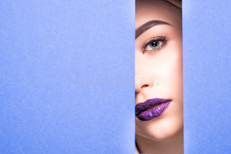 Face of young beautiful girl with a bright make-up and violet lips looks through a hole in violet paper. Closeup beauty portrait stock image Face of young beautiful girl with a bright make-up and violet lips looks through a hole in violet paper. Closeup beauty portrait stock image