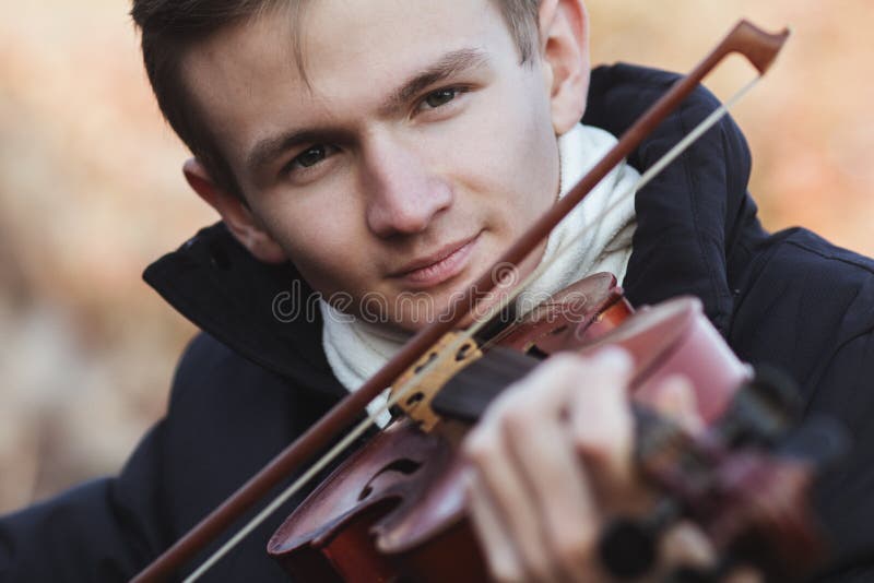 Face of a young elegant man playing the violin on autumn nature backgroung, a boy with a bowed instrument practicing, musical performance outdoors, concept of hobby and art