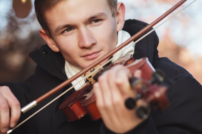 Face of a young elegant man playing the violin on autumn nature backgroung, a boy with a bowed instrument practicing, musical performance outdoors, concept of hobby and art