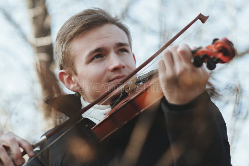 Face of a young elegant man playing the violin on autumn nature backgroung, a boy with a bowed instrument practicing, musical performance outdoors, concept of hobby and art