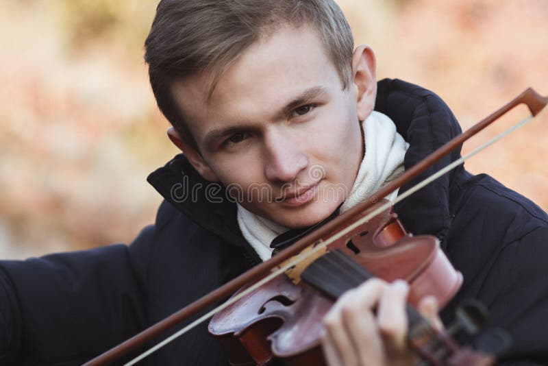 Face of a young elegant man playing the violin on autumn nature backgroung, a boy with a bowed instrument practicing, musical performance outdoors, concept of hobby and art