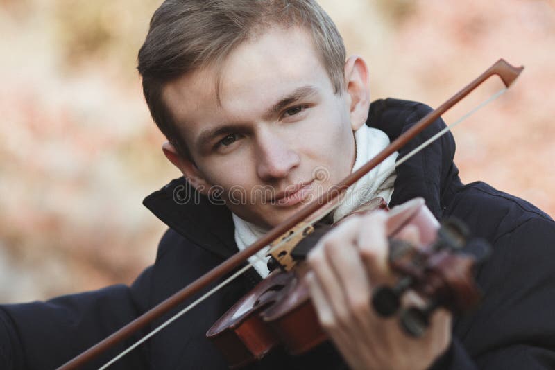 Face of a young elegant man playing the violin on autumn nature backgroung, a boy with a bowed instrument practicing, musical performance outdoors, concept of hobby and art