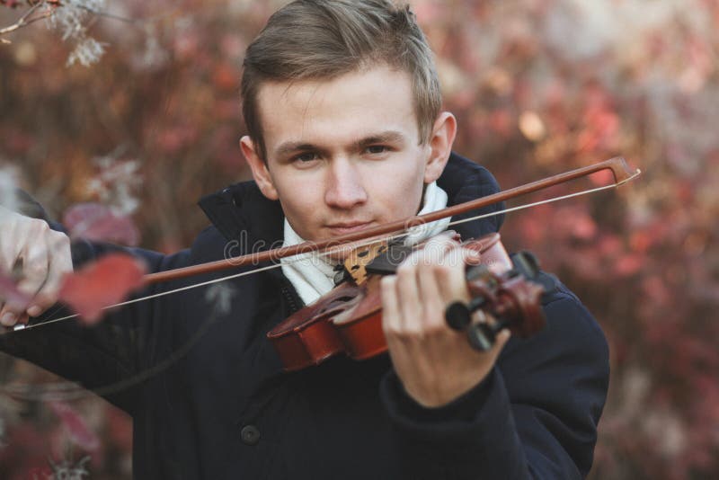 Face of a young elegant man playing the violin on autumn nature backgroung and confidently looking straight, boy with musical instrument practicing, performance outdoors, concept of hobby and art