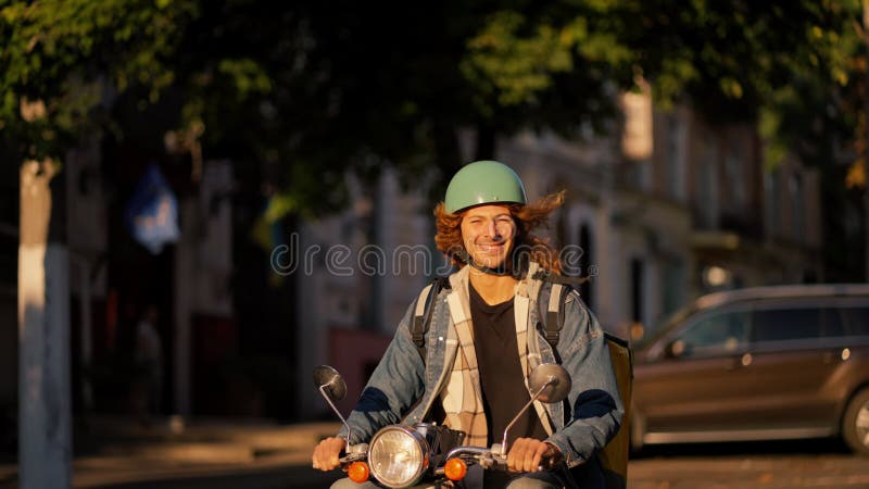Facet z długimi włosami kręconymi w dżinsowej kurtce na zielonych motorowerach wzdłuż drogi w pięknym letnim mieście