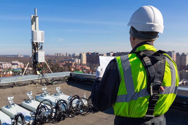 Professional industrial climber in helmet and uniform reads technical documentation and antennas of GSM DCS UMTS LTE bands, outdoor radio units on the roof. Working process of upgrading telecommunication equipment. Professional industrial climber in helmet and uniform reads technical documentation and antennas of GSM DCS UMTS LTE bands, outdoor radio units on the roof. Working process of upgrading telecommunication equipment