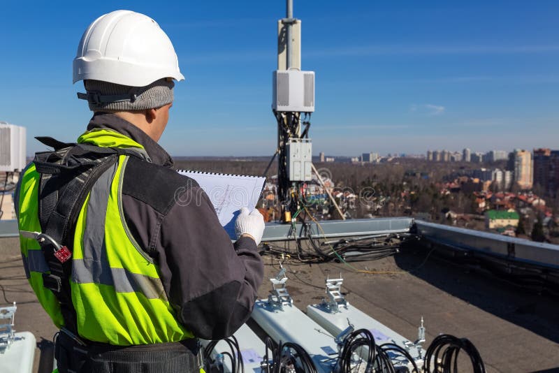 Professional industrial climber in helmet and uniform reads technical documentation and antennas of GSM DCS UMTS LTE bands, outdoor radio units on the roof. Working process of upgrading telecommunication equipment. Professional industrial climber in helmet and uniform reads technical documentation and antennas of GSM DCS UMTS LTE bands, outdoor radio units on the roof. Working process of upgrading telecommunication equipment