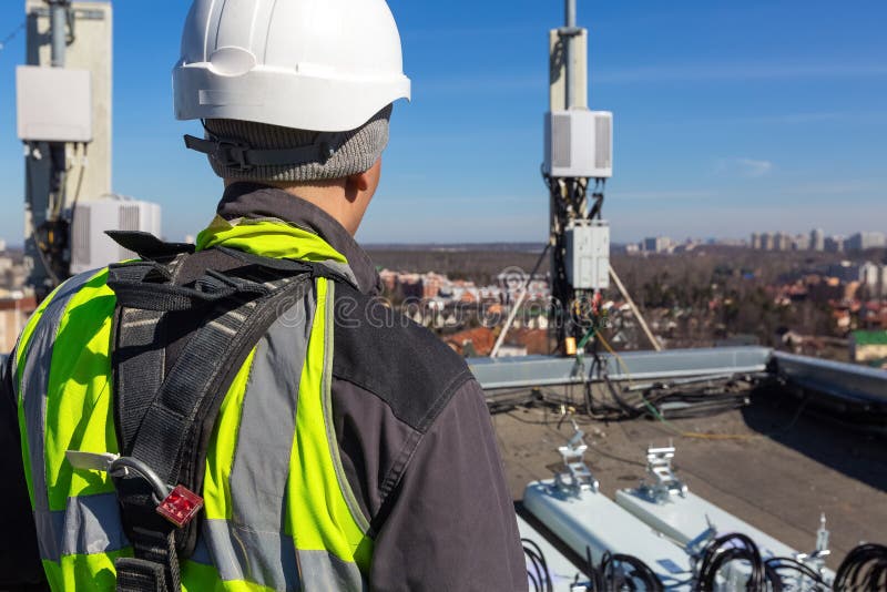 Professional industrial climber in helmet and uniform  and  antennas of GSM  DCS UMTS LTE bands, outdoor radio units  on the roof. Working process of upgrading telecommunication equipment. Professional industrial climber in helmet and uniform  and  antennas of GSM  DCS UMTS LTE bands, outdoor radio units  on the roof. Working process of upgrading telecommunication equipment