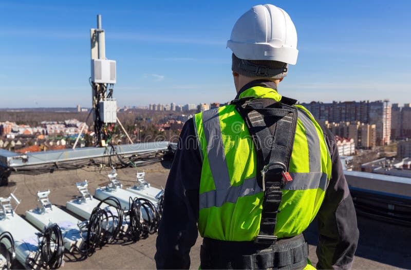 Professional industrial climber in helmet and uniform  and  antennas of GSM  DCS UMTS LTE bands, outdoor radio units  on the roof. Working process of upgrading telecommunication equipment. Professional industrial climber in helmet and uniform  and  antennas of GSM  DCS UMTS LTE bands, outdoor radio units  on the roof. Working process of upgrading telecommunication equipment