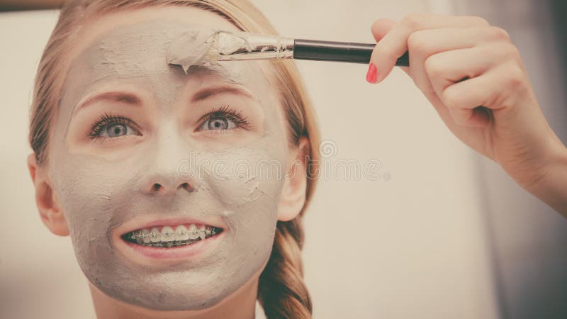 Facial dry skin and body care, complexion treatment at home concept. Happy young woman applying grey mud mask on her face with brush. Facial dry skin and body care, complexion treatment at home concept. Happy young woman applying grey mud mask on her face with brush