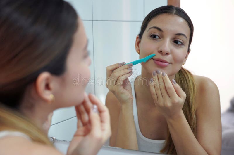 Facial hair removal. Close-up of beautiful young woman shaving her face by razor at home. Pretty girl using razor on bathroom