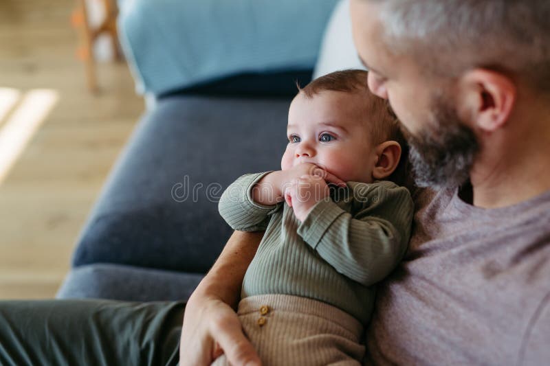 Fader som håller babypojken i armar som sitter med honom på soffan. arkivfoto