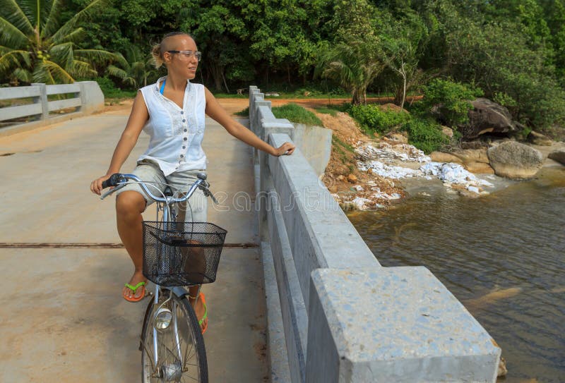 Fahrrad der jungen Frau Reitüber Flussbrücke nahe bei tropischem Park lizenzfreies stockfoto Fahrrad der jungen Frau Reitüber Flussbrücke nahe bei tropischem Park lizenzfreies stockfoto