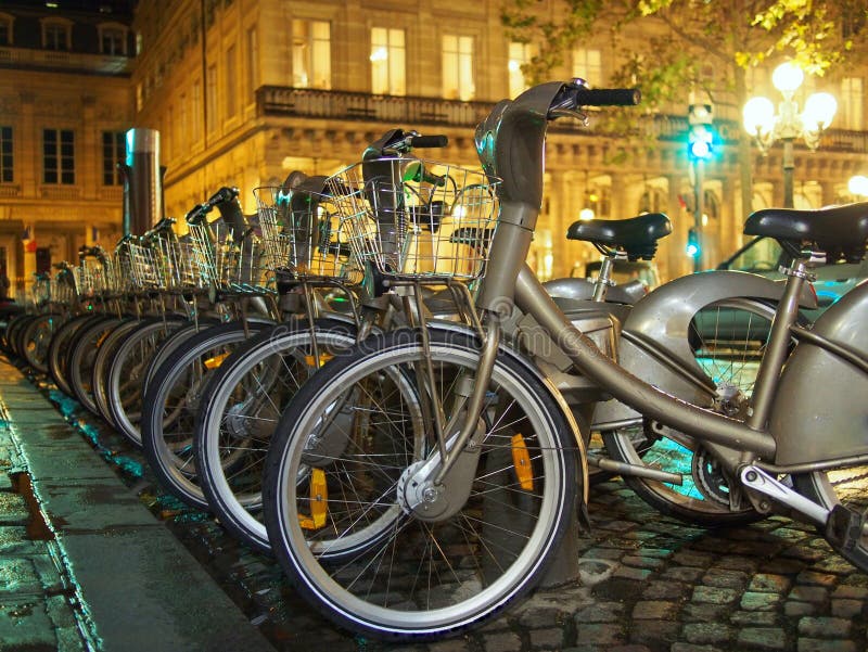 Bicycles in lines waiting to be picked at the station for a romantic ride in Paris city by night. Public Velib bicycle sharing system, with brands removed. Typical street pavement, lights and buildings behind. Bicycles in lines waiting to be picked at the station for a romantic ride in Paris city by night. Public Velib bicycle sharing system, with brands removed. Typical street pavement, lights and buildings behind.