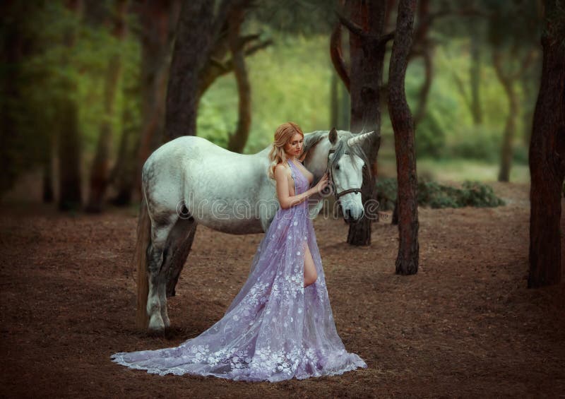 A fairy in a purple, transparent dress with a long train - caught a unicorn. Fantastic magical, radiant horse. Blonde girl walking with pegasus in the forest. Artistic Photography