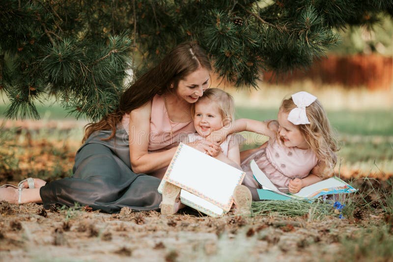 Fam?lia feliz - a mam? e duas filhas est?o sentando-se em um prado e est?o lendo-se um livro Piquenique imagem de stock Fam?lia feliz - a mam? e duas filhas est?o sentando-se em um prado e est?o lendo-se um livro Piquenique imagem de stock