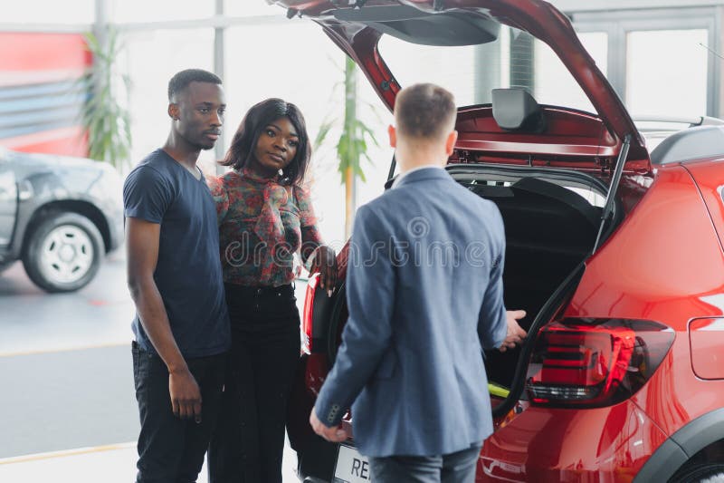 Famiglia afro-americana al concessionario d'auto. il venditore mostra il bagagliaio di una nuova auto rossa immagine stock libera da diritti