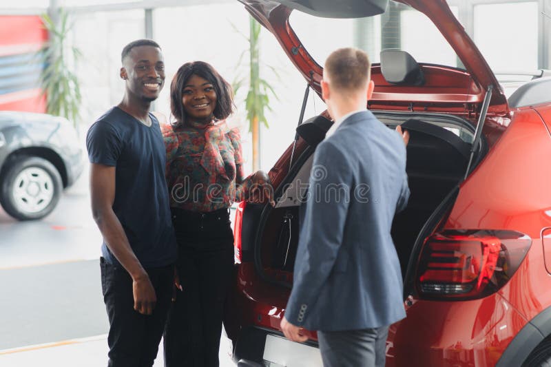 Famiglia afro-americana al concessionario d'auto. il venditore mostra il bagagliaio di una nuova auto rossa fotografie stock libere da diritti