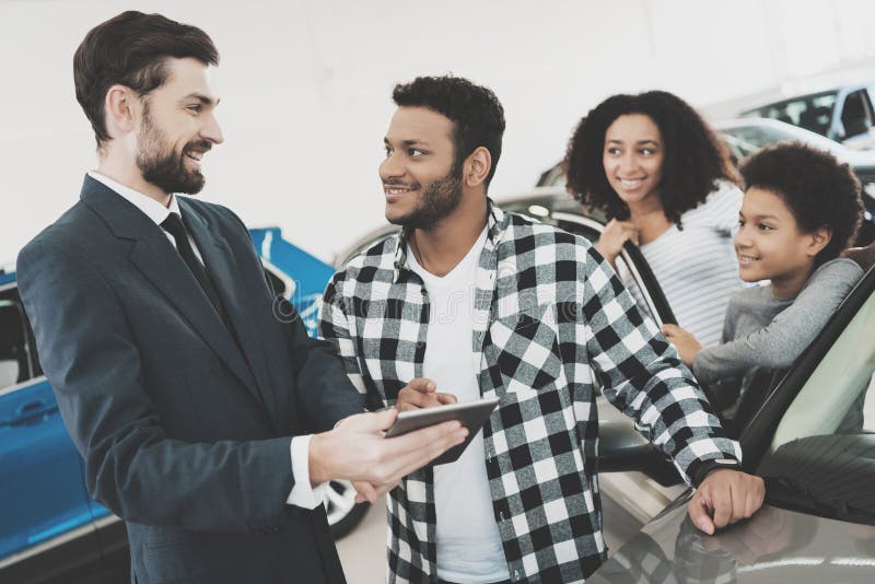 Famiglia afroamericana al concessionario auto Il rappresentante sta mostrando i dettagli per la nuova automobile sulla compressa fotografie stock