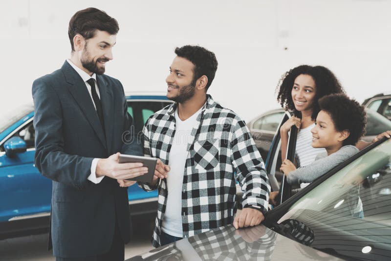 Famiglia afroamericana al concessionario auto Il rappresentante sta mostrando i dettagli per la nuova automobile sulla compressa fotografia stock