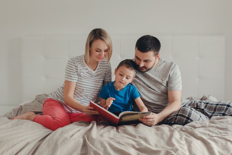 Famiglia di tre persone che si siede sul letto in libro di lettura della camera da letto immagini stock Famiglia di tre persone che si siede sul letto in libro di lettura della camera da letto immagini stock