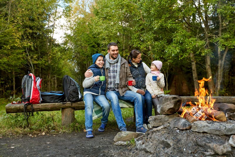 Family, camping and tourism concept - happy mother, father, son and daughter sitting on bench and drinking hot tea from cups at camp fire in woods. Family, camping and tourism concept - happy mother, father, son and daughter sitting on bench and drinking hot tea from cups at camp fire in woods