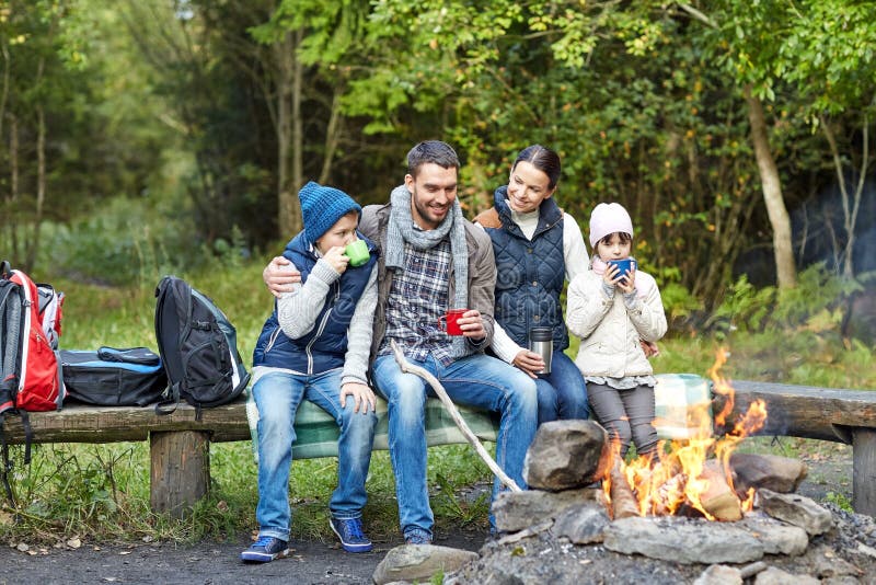 Camping, travel, tourism, hike and people concept - happy family sitting on bench and drinking hot tea from cups at camp fire in woods. Camping, travel, tourism, hike and people concept - happy family sitting on bench and drinking hot tea from cups at camp fire in woods