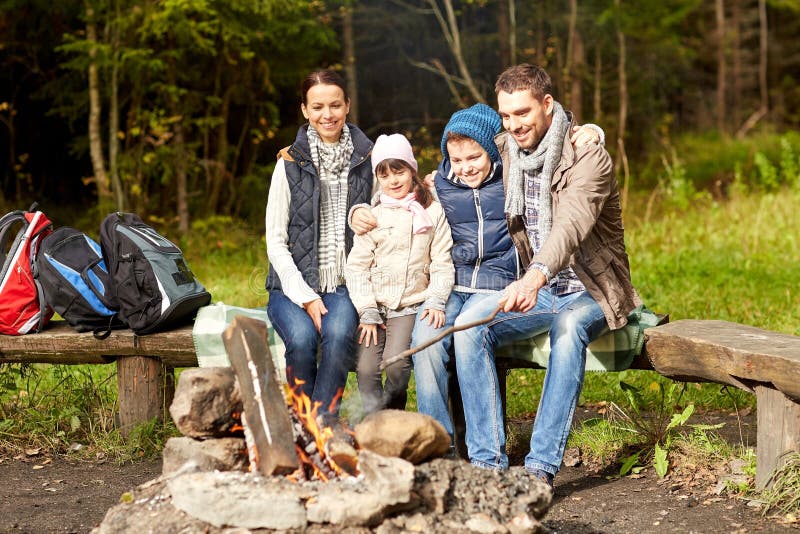 Camping, travel, tourism, hike and people concept - happy family sitting on bench at camp fire in woods. Camping, travel, tourism, hike and people concept - happy family sitting on bench at camp fire in woods