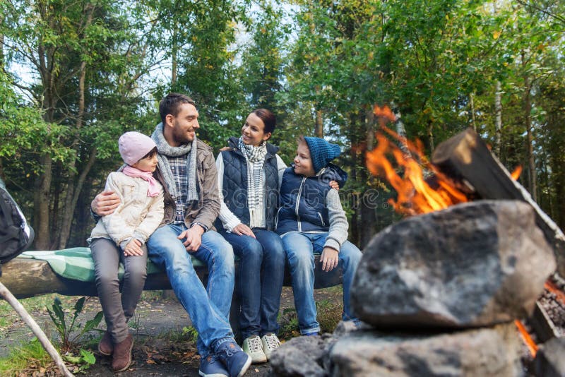 Camping, travel, tourism, hike and people concept - happy family sitting on bench and talking at camp near campfire in woods. Camping, travel, tourism, hike and people concept - happy family sitting on bench and talking at camp near campfire in woods