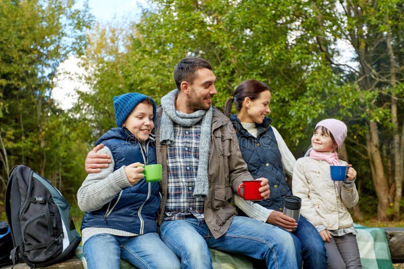 Family, camping and tourism concept - happy mother, father, son and daughter sitting on bench and drinking hot tea from cups at camp in woods. Family, camping and tourism concept - happy mother, father, son and daughter sitting on bench and drinking hot tea from cups at camp in woods