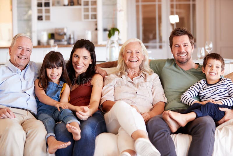 Familia blanca feliz de tres generaciones que se sienta en un sofá junto en casa que sonríe a la cámara, vista delantera imagen de archivo Familia blanca feliz de tres generaciones que se sienta en un sofá junto en casa que sonríe a la cámara, vista delantera imagen de archivo