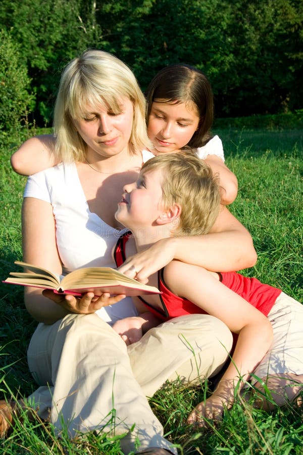 The young woman sits on a grass and reads the book to the children. The young woman sits on a grass and reads the book to the children