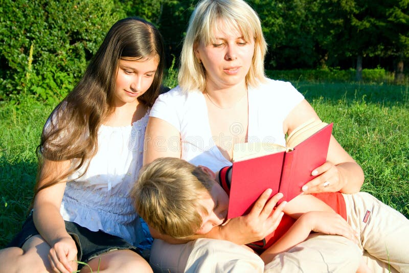 The young woman sits on a grass and reads the book to the children. The young woman sits on a grass and reads the book to the children