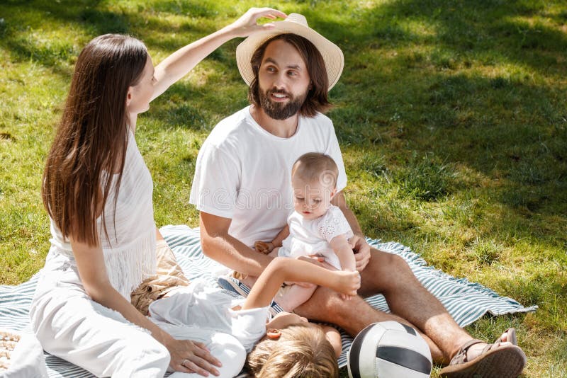 Familia joven feliz en una comida campestre soleada del verano Están llevando la ropa blanca y se están sentando con los niños en imágenes de archivo libres de regalías Familia joven feliz en una comida campestre soleada del verano Están llevando la ropa blanca y se están sentando con los niños en imágenes de archivo libres de regalías