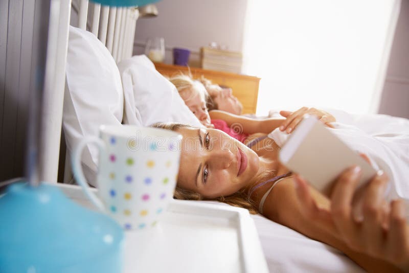 Familj i säng som moderräckvidder som kontrollerar mobiltelefonen arkivbild Familj i säng som moderräckvidder som kontrollerar mobiltelefonen arkivbild