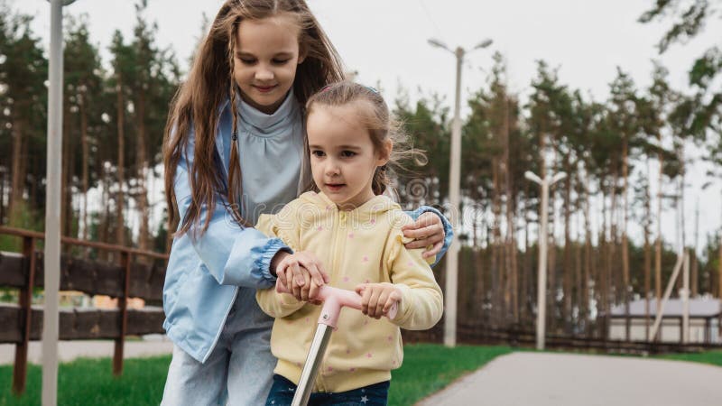 Familjevårdskoncept. En liten flicka lär sin syster att åka med en skoter i parken royaltyfri bild