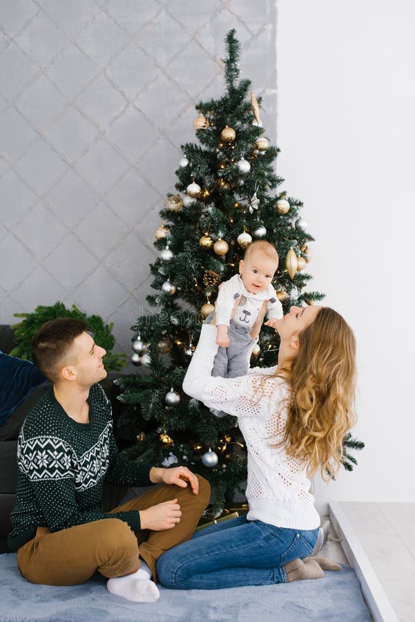 Friendly young family: dad, mom and baby sitting near the Christmas tree. Mom lifted the baby up in her arms and smiles at him, dad looks at them. Friendly young family: dad, mom and baby sitting near the Christmas tree. Mom lifted the baby up in her arms and smiles at him, dad looks at them