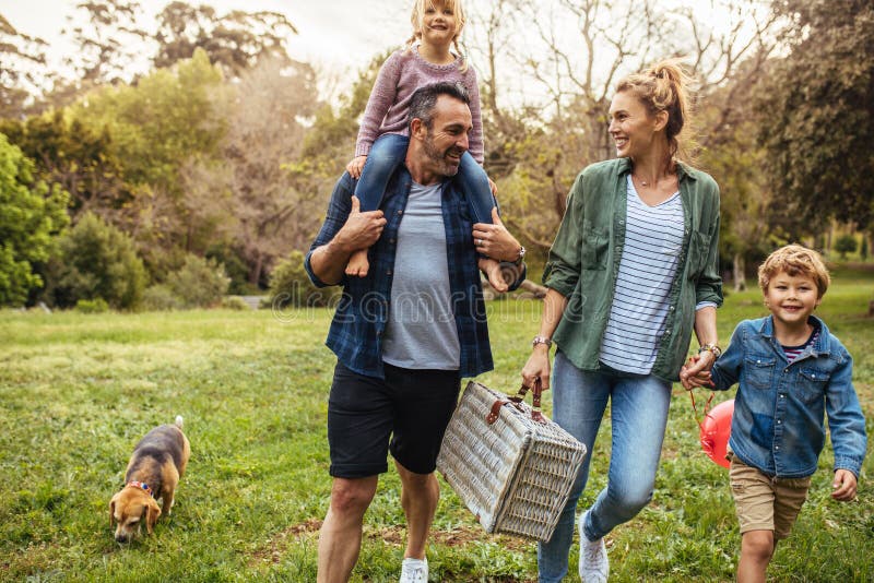 Happy family with dog walking towards the picnic spot in the garden. Man carrying his daughter on shoulders with women holding son hand and carrying a picnic basket. Happy family with dog walking towards the picnic spot in the garden. Man carrying his daughter on shoulders with women holding son hand and carrying a picnic basket