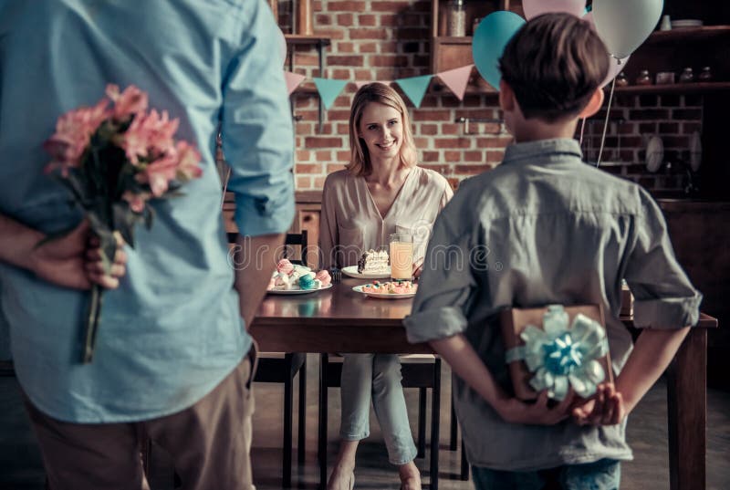 Family celebrating mother`s birthday in decorated kitchen. Husband with flowers, son with a gift box, mother is smiling waiting for presents. Family celebrating mother`s birthday in decorated kitchen. Husband with flowers, son with a gift box, mother is smiling waiting for presents