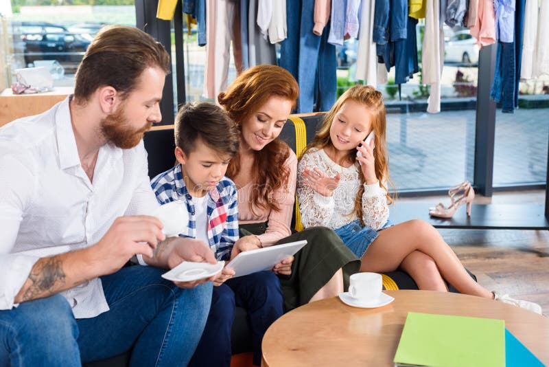 family drinking coffee and using digital tablet while resting on sofa in boutique