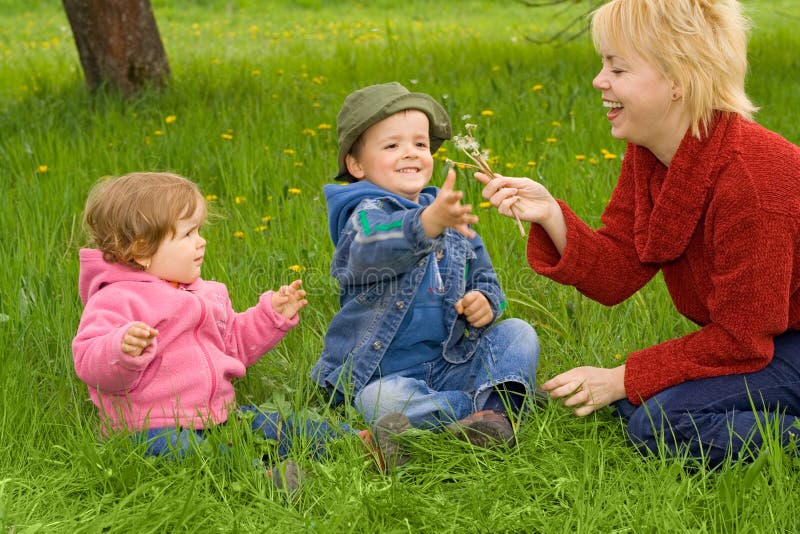 Young woman and children blowing dandelions sitting in the grass in springtime. Young woman and children blowing dandelions sitting in the grass in springtime