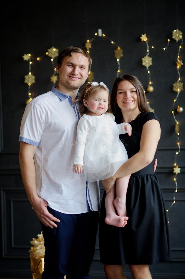 Family with little daughter is photographed in day of a holiday. Room is festively decorated. Baby is dressed in a white dress, on the head of her a wreath. Girlie is capricious. Parents smile.