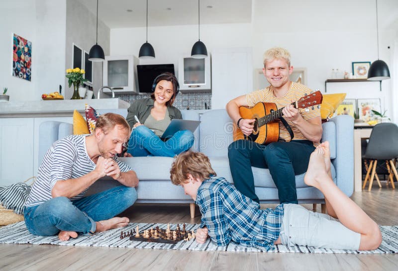 Family at living room together. Father playing chess with little son, mother learning online using laptop, older son playing stock photos Family at living room together. Father playing chess with little son, mother learning online using laptop, older son playing stock photos