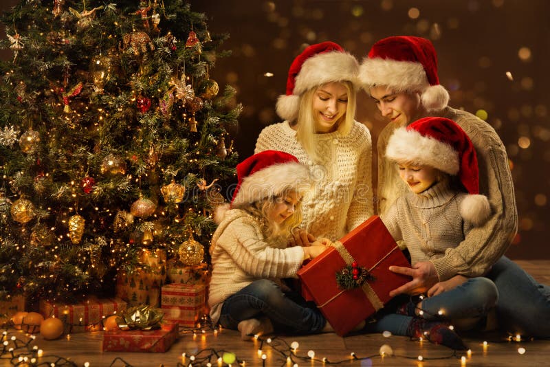 Family open Christmas Present Gift Box next to Decorated Xmas Tree, Happy Parents with Children in cozy Dark Holiday Room Interior