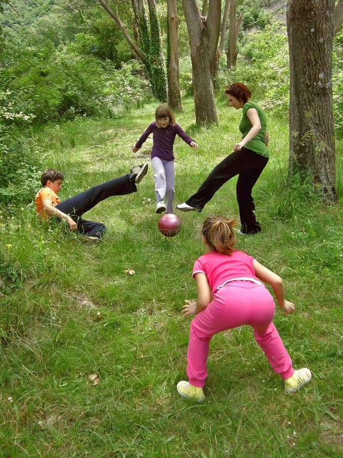 Four people play sports. Mom with three children (best friends) boy with two sisters play soccer - one girl kicking a red ball football in nature on green grass. Vertical color photo. Four people play sports. Mom with three children (best friends) boy with two sisters play soccer - one girl kicking a red ball football in nature on green grass. Vertical color photo.