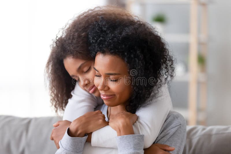 Family sitting on couch in living room at home african teenager daughter embracing from behind beloved mother. Relative people, older younger sisters, new mom for adopted child, love and care concept. Family sitting on couch in living room at home african teenager daughter embracing from behind beloved mother. Relative people, older younger sisters, new mom for adopted child, love and care concept