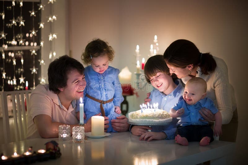 Young happy family with three kids - teenager son, toddler girl and baby boy - celebrating the birthday of their son blowing out the candles on a cake in a dark living room. Young happy family with three kids - teenager son, toddler girl and baby boy - celebrating the birthday of their son blowing out the candles on a cake in a dark living room