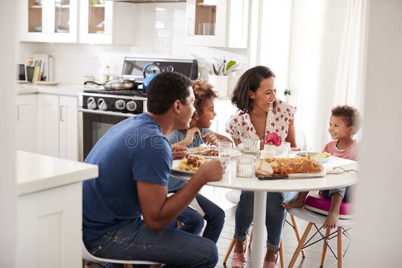 Família afro-americano nova que come uma refeição junto na tabela em sua cozinha, vista da entrada