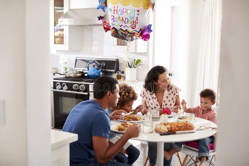 Família afro-americano nova que tem uma refeição do aniversário junto na tabela em sua cozinha, vista da entrada