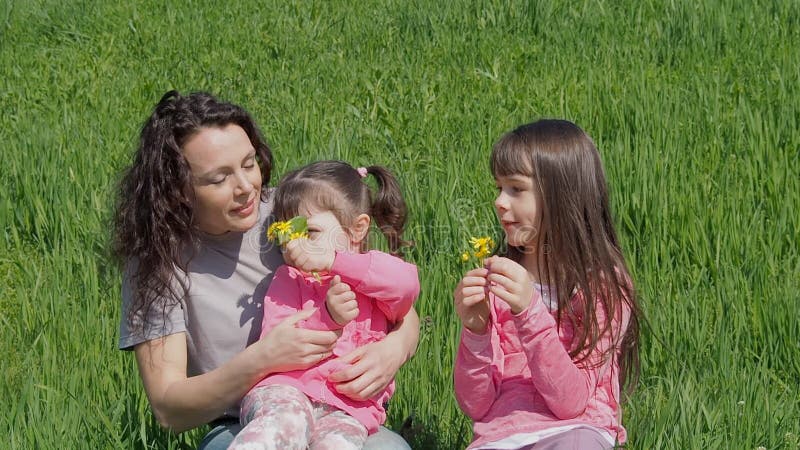 A família na grama verde está aspirando flores A mamã e a filha estão sentando-se no gramado Mulher com as meninas na natureza de